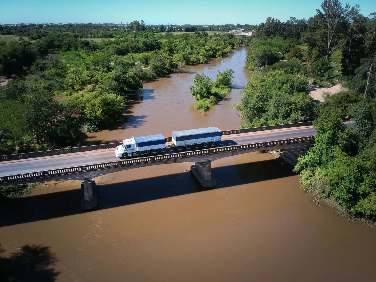 Camión con semirremolque cruzando un puente sobre un río de agua marrón, entre monte verde del litoral.