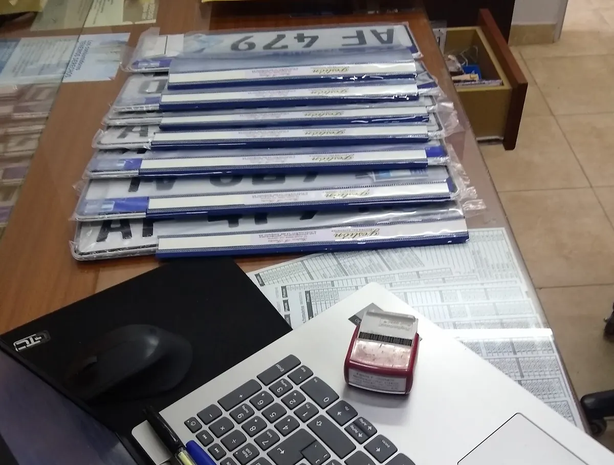 Placas patente nuevas, todavía en su envoltorio, apiladas sobre el escritorio de la oficina junto a una notebook y el sello.